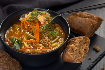 Tasty soup with pieces of beef and vegetables in black bowl served with spices and breads on dark wooden background.