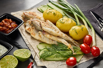 Fried smelts serving with potatoes, vegetables, lime and sauces. Grey slate background. Top view