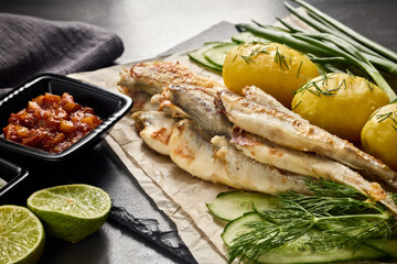 Fried smelts serving with potatoes, vegetables, lime and sauces. Grey slate background. Selective focus on fish.