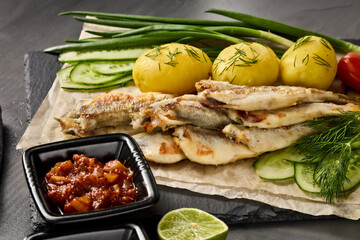 Fried smelts serving with potatoes, vegetables, lime and sauces. Grey slate background.
