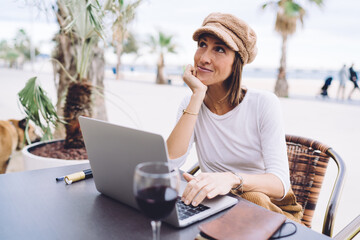 Dreamy young female in trendy wear pondering on idea for vlog working in social networks via laptop computer, positive woman digital nomad satisfied with freelance distance job on cafe terrace