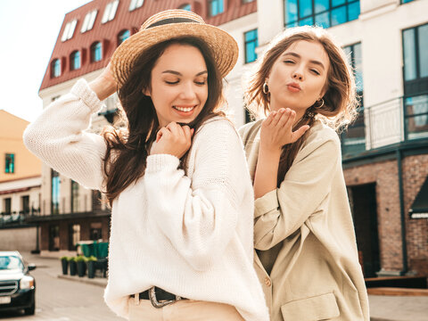 Two young beautiful smiling hipster female in trendy white sweater and coat. Sexy carefree women posing on the street background in hats. Positive models having fun outdoors. Hugging