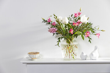 A bouquet of white tulips, pink eustoma, hyacinth, eucalyptus in fluted glass vase on panel of an artificial fireplace. Tea in a cup, faience figurines of a bird and an angel. Empty space.