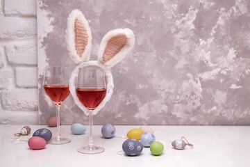 Obraz na płótnie Canvas Two glasses of rose or red wine with bunny ears and Easter decorations, colorful eggs on white table, on bright background. Copy space for text.