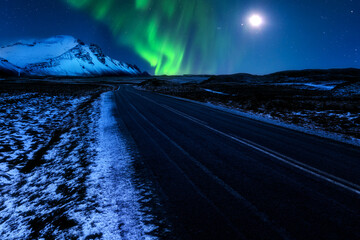 Icelandic Hringvegur ring-road (route 1) leads towards the snow-covered Vestrahorn mountains, below the Northern Lights and the moon. - Powered by Adobe