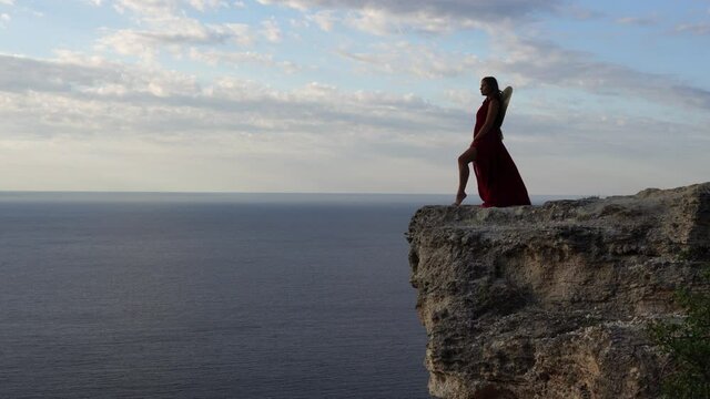 A young woman dressed in a red dress stands next to the sea on a rock, A girl enjoys the view of the sea and the sunrise, her hair is blown by the wind, the hem of the dress is fluttering.