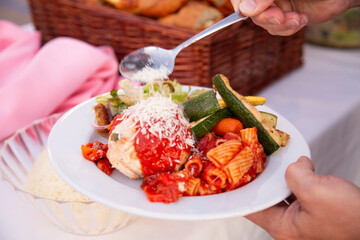 Hand holding spoon sprinkling Parmesan cheese on pasta and chicken dinner with vegetables at buffet table