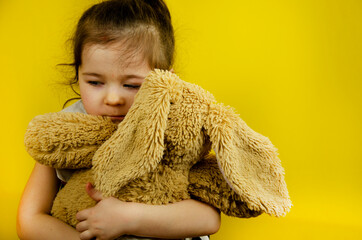 Little girl with a teddy bear. Little girl hugs a soft toy looks up. The child hugs his toy. The child plays alone. Girl with a toy on a yellow background. The concept of parental inattention to child