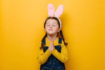 Obraz na płótnie Canvas Close up portrait of beautiful cute preschool kid girl in pink bunny fluffy ears is praying for something, dressed in sweater, isolated on yellow studio background. Easter holiday and wish concept