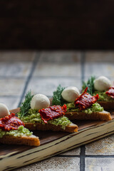 Mini triangle canapes bruschetta with avocado, mozarella and dried tomatos on the rustic board on the vintage tiled background