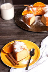 Concepto de un plato amarillo con un trozo de pastel con azúcar glash, ramas de canela, una rodaja de naranja y una cuchara de postre sobre un soporte de madera gris.