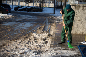 A manual worker sweeps the remnants of snow off a wet pavement with a broom