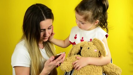 The child asks his mother to play with him. Social media addiction. Busy mom at home. Baby and mom on a yellow background. Deficit of parental attention to children. Unattended child with a soft toy. 