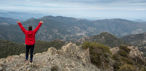 Back view of a backpacker woman with raised arms standing on rocky top enjoying mountain range panorama.