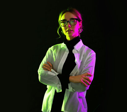 Portrait young woman in glasses pink and green light on a dark background.