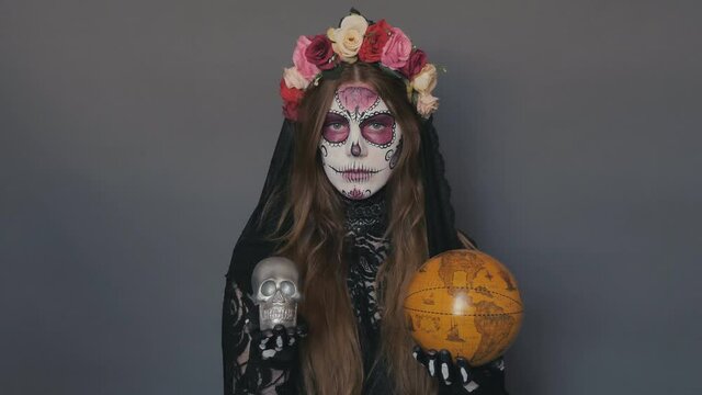 woman in the image of death Santa Muerte or sugar mexican skull with a globe and a skull in her hands on a gray background. halloween make-up