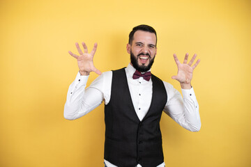 Young man with beard wearing bow tie and vest smiling funny doing claw gesture as cat, aggressive...