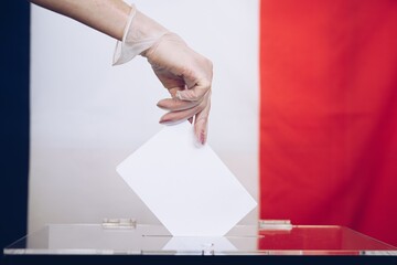 Woman's hand in medical glove throwing her vote into the ballot box. Flag France in background.