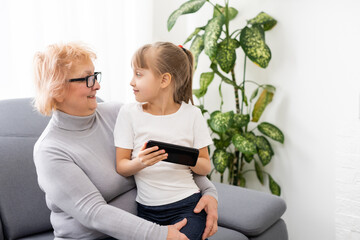 Happy grandmother and granddaughter using phone together, sitting on cozy sofa at home, browsing mobile device apps, grandma with grandchild playing game, looking at screen, having fun