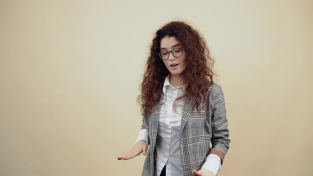 The cheerful young woman dances with open palms, waving her hands left and right, humming the music. Cretaceous in gray jacket and white shirt, with glasses posing isolated on a beige background in
