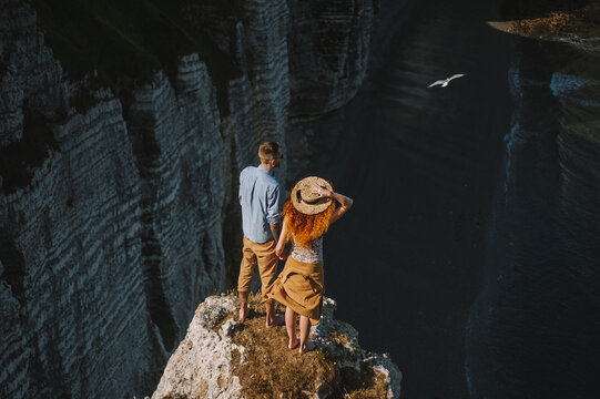 a guy and a red-haired girl in a straw hat stand in summer in sunny weather on the edge of a beautiful cliff and look at the seashore back view