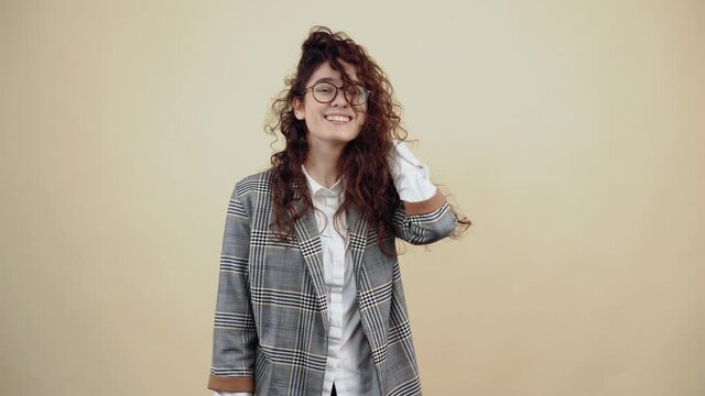 The young woman with a wide, shy smile puts her hand in her hair and smiles wide. Cretaceous in gray jacket and white shirt, with glasses posing isolated on a beige background in the studio. The