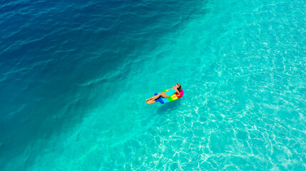 AERIAL. Young Woman weared in blue swim suit relaxing on a colourful air mattress. View from a drone to young traveller swimming in to the blue sea. Travel concept.