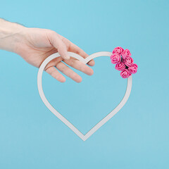 Hand holding white heart frame decorated with pink roses. Pastel blue background. Minimal love concept.