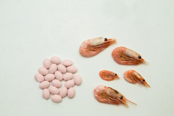 Isolated on a white background 5 shrimps of different sizes from large to small lie next to pink pills. The tablets contain vitamins and minerals from seafood.