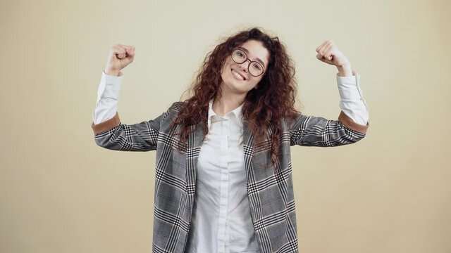 Strong and confident young woman, with her hands on her side up, inspires strength, she shows her muscles. Cretaceous in gray jacket and white shirt, with glasses posing isolated on a beige background
