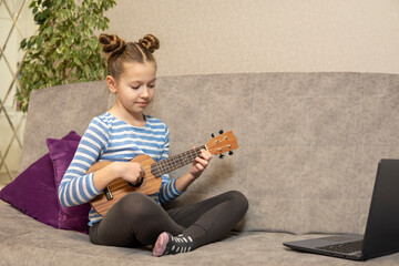 Teenage girl learns to play the ukulele and looks into her laptop.