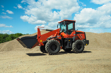 Obraz premium A red-black front-end loader with small wheels against the background of a large pile of stone sand and a blue sky with white clouds. Side view.