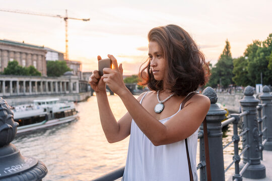 Female student taking photograph with smartphone by river, Berlin, Germany