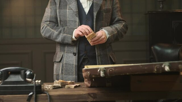 Mafia gangster in plaid suit in 1920 stands at table in room and counts old money. The concept of dividing the loot after bank or shop robbery.