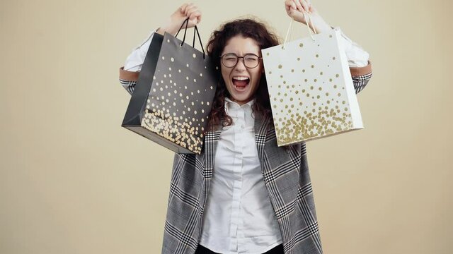 The happy young woman holds two cardboard bags in me, satisfied with the purchases made, she smiles with her teeth out. Cretaceous in gray jacket and white shirt, with glasses posing isolated on a