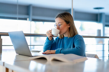 Female higher education student looking at laptop and drinking coffee in university cafe