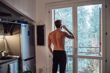 Mid adult man with tattoos drinking coffee and looking out through kitchen window