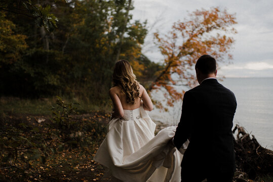 Romantic young groom carrying bride's wedding dress train on lakeside, rear view