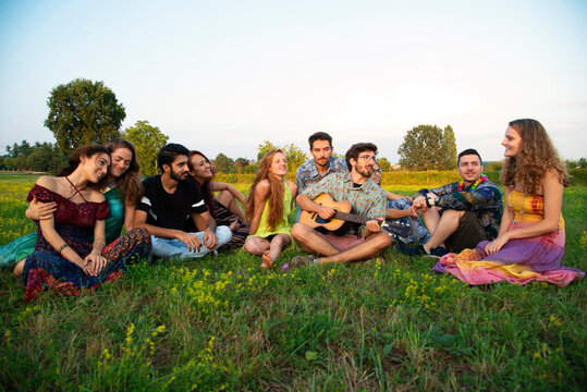 Large group of young adults sitting in field listening to acoustic guitar