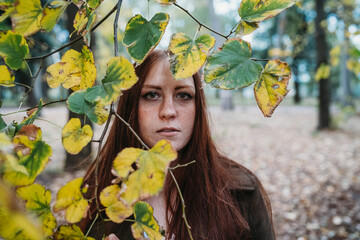 Young woman with long red hair behind twig of autumn leaves in park, portrait