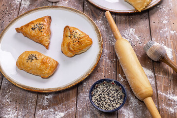 Chebureks on a plate, sauce, flour, sesame seeds, rolling pin on a brown wooden background.