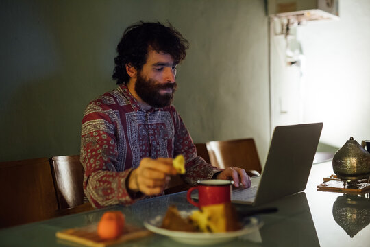 Bearded young man having breakfast, using laptop at home