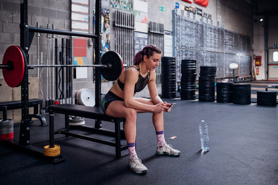 Young woman training, sitting on weights bench in gym looking at smartphone