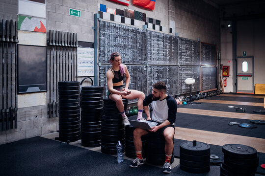 Young woman and man training together in gym, sitting on weights with smartphone and laptop