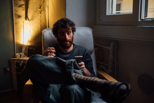 Bearded young man smoking, using smartphone at home