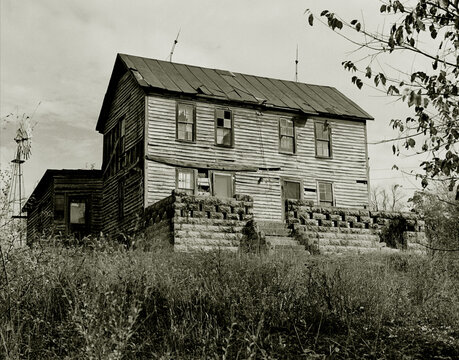 House On The Hill. A Sepia Toned Black And White Vintage Image Of An Abandoned House On A Hilltop With A Windmill Out Back In Rural Northern Misouri. Yarrow, Missouri, USA, 1978.