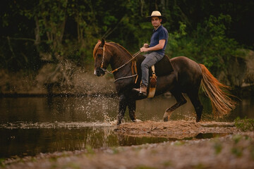 White man riding a horse inside a river, wearing a straw hat, with his horse in a saddle and...