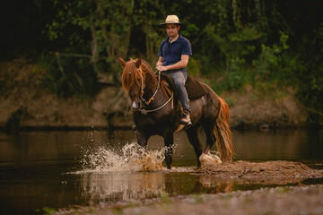 White man riding a horse inside a river, wearing a straw hat, with his horse in a saddle and...