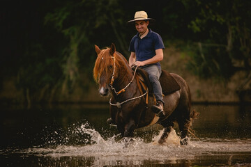 White man riding a horse inside a river, wearing a straw hat, with his horse in a saddle and...