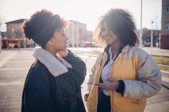 Two Cool Young Female Friends Sharing Earphones On Urban Sidewalk